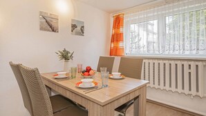 A dining area with a wooden table, chairs, and a window with curtains. There are wall-mounted pictures and a radiator. - Cozy vacation apartment in Zinnowitz with garden, only 750 m from the beach (Zinnowitz)