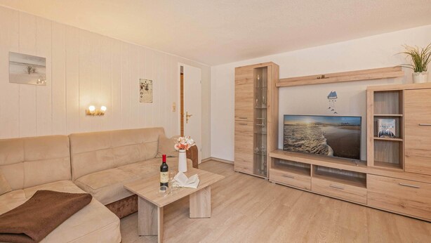 A living room with a beige sofa, a coffee table with a wine bottle and glasses, a wooden cabinet with a glass display, and a wall-mounted TV. - Cozy vacation apartment in Zinnowitz with garden, only 750 m from the beach (Zinnowitz)