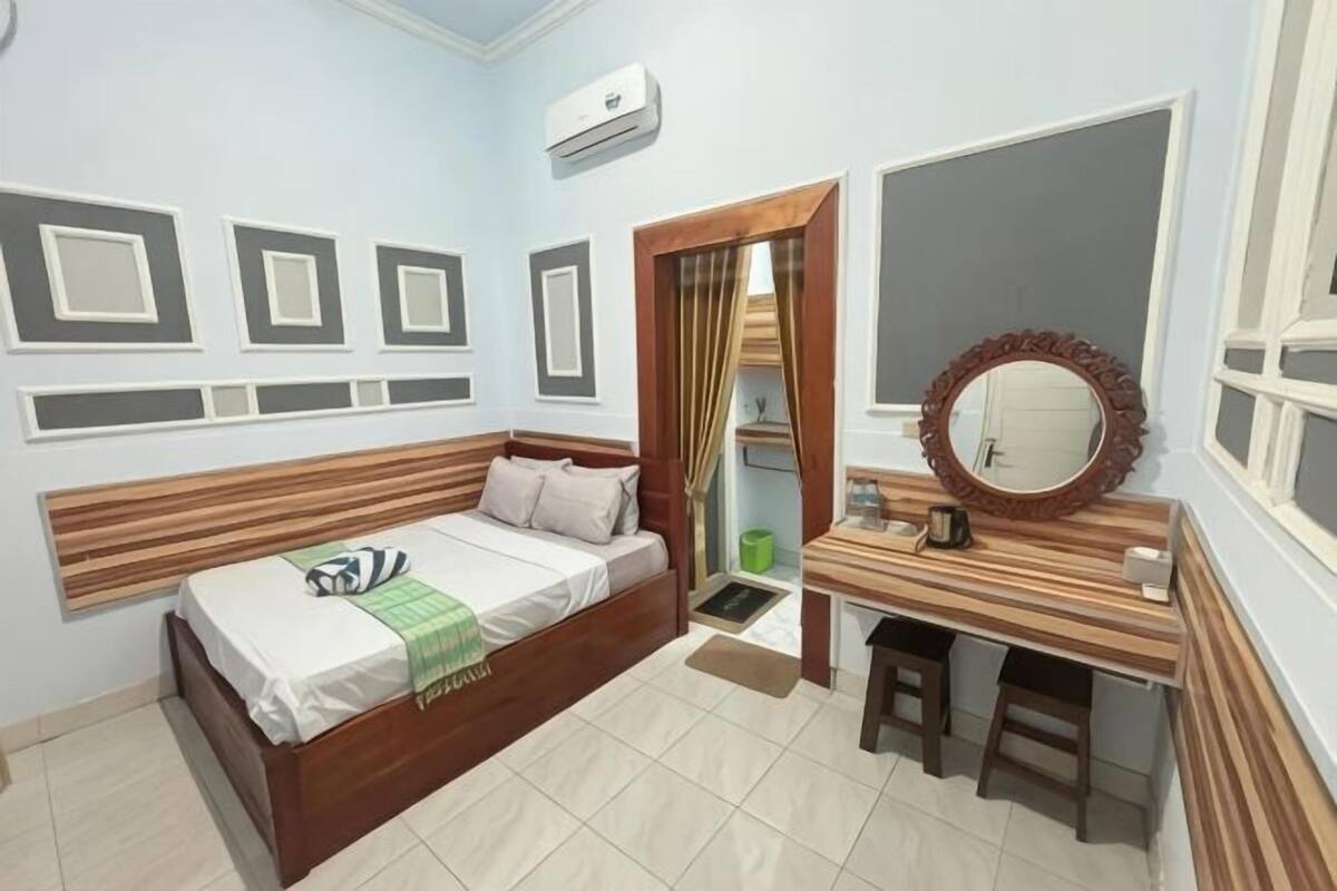 A bedroom with a bed, a wooden desk, a mirror, and a window.
