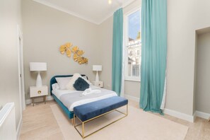 A bedroom with a bed, bedside tables, a window with curtains, a wall decoration, and a view of buildings outside. - Stunning Notting Hill Apartment (PS4) (Greater London)