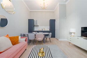 A modern living room with a pink sofa, a dining area with chairs, a kitchen with a stove, and a TV on a white cabinet. - Stunning Notting Hill Apartment (PS4) (Greater London)