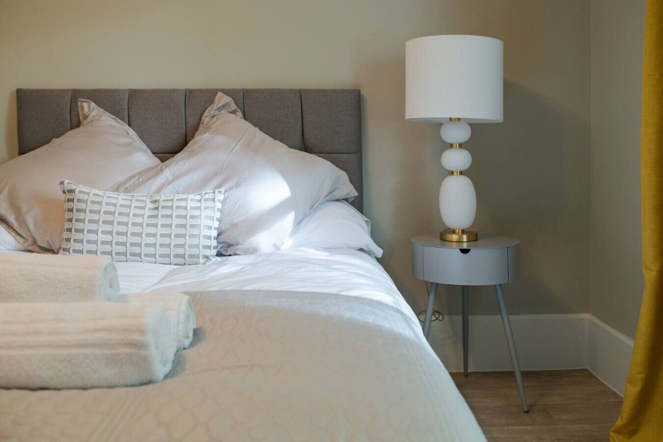 Una cama bien hecha con sábanas blancas, cabecero gris y mesita de noche con lámpara.
