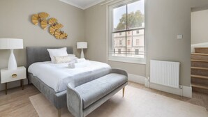 Ein modernes Schlafzimmer mit einem großen Bett, Nachttischlampen, einer Bank, einem Heizkörper und einem Fenster mit Blick auf Gebäude.