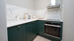 Eine Küche mit dunkelgrünen Schränken, einer weißen Marmor-Spritzschutzwand, einer Edelstahl-Dunstabzugshaube, einem eingebauten Ofen und einer weißen Arbeitsplatte mit goldenem Wasserhahn und einer Kaffeemaschine.