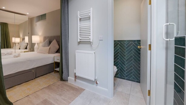 A bedroom with a bed, bedside lamps, a radiator, and a bathroom with a herringbone tile backsplash. - Stylish Hyde Park Apartment (PS2) (Greater London)
