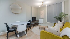 A modern living room with a round dining table, two chairs, a yellow sofa, a TV, and a large window with blinds. - Trendy Flat by Hyde Park (PS1) (Greater London)