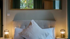 Een hotelkamer met een bed, twee nachtlampjes, een hoofdbord, een raam met gordijnen en een spiegel aan de muur.