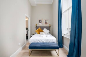 A bedroom with a bed, a bench, a window with curtains, and a door leading to a bathroom.