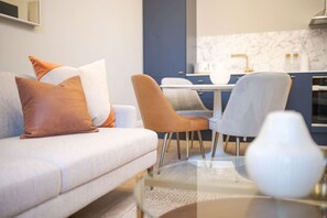 A modern living room with a sofa, two chairs, a glass coffee table, and a kitchen in the background.