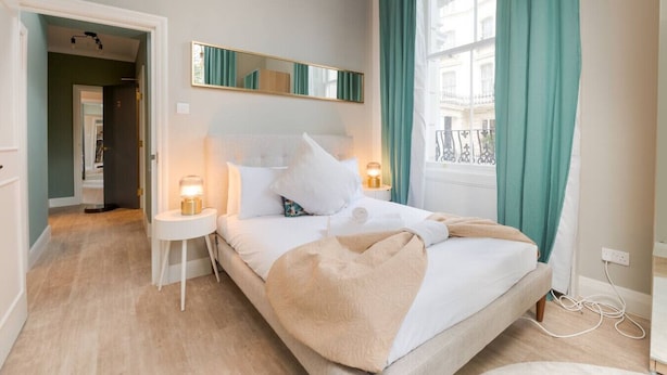 Een slaapkamer met een bed, bedlampen, een spiegel en een raam met gordijnen.