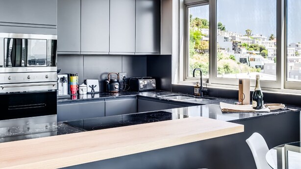 A modern kitchen with a central island, black cabinets, and a view of a cityscape through large windows. - Deluxe Urban Retreat Summit on Quarter (Cape Town)