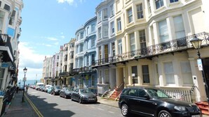 Une rangée d’immeubles à plusieurs étages dotés de balcons, des voitures garées et un ciel dégagé.