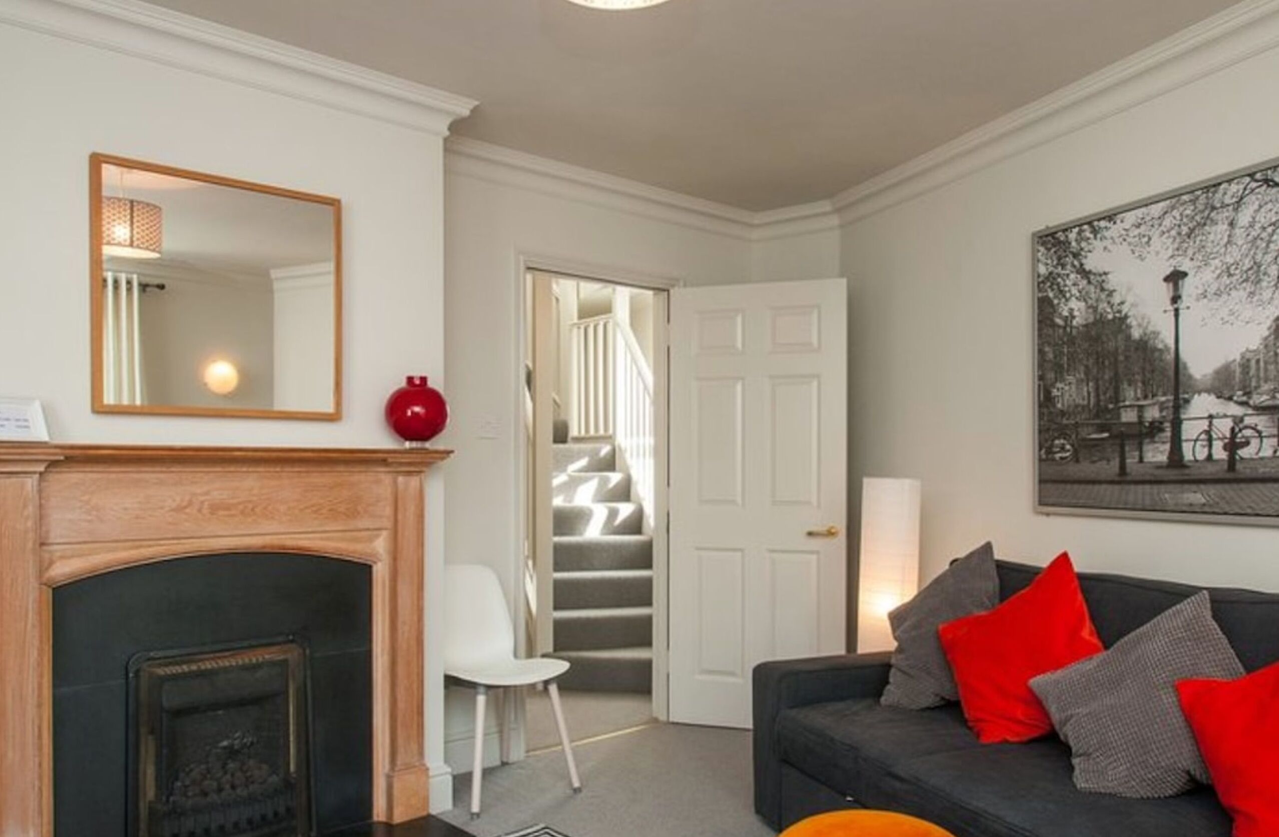 A living room with a fireplace, a sofa with red and grey cushions, a white chair, a large mirror, and a staircase leading to another room.