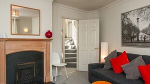 A living room with a fireplace, a sofa with red and grey cushions, a white chair, a large mirror, and a staircase leading to another room.