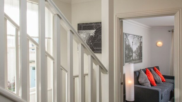 A staircase leading to a room with a grey sofa and red cushions, a black and white framed picture on the wall, and a window with white blinds.