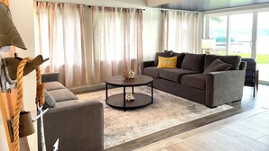 Un salon moderne avec un ventilateur de plafond, un canapé gris agrémenté d’un coussin jaune, une table basse ronde et une grande fenêtre avec des rideaux.