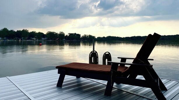 Une terrasse en bois avec un fauteuil de relaxation et une table, donnant sur un lac paisible dont on aperçoit la rive au loin.