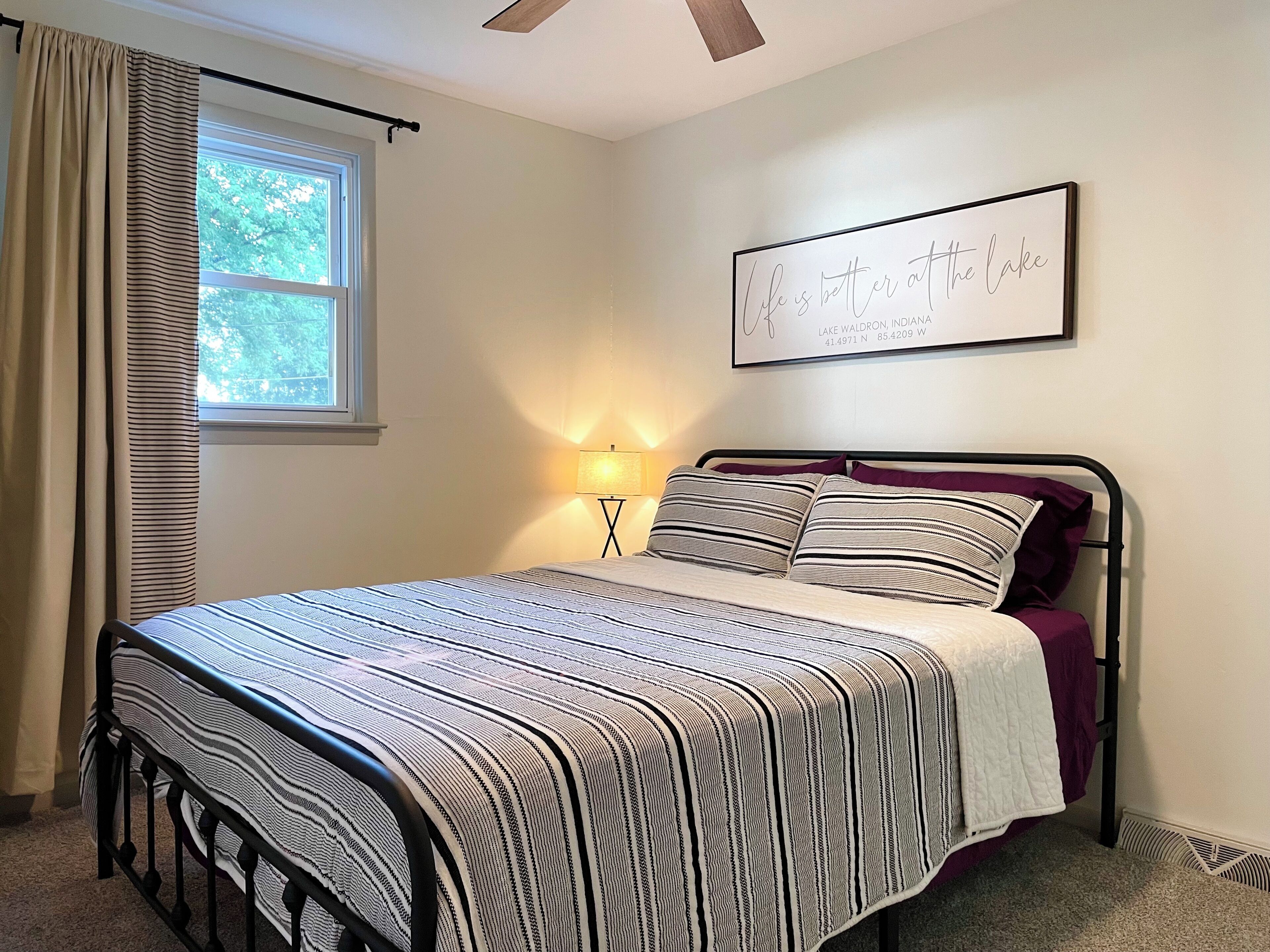 A bedroom with a bed, a nightstand, a lamp, a window with curtains, and a wall with a framed quote.