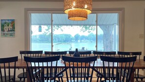 Un espace repas avec une grande table et des chaises, un luminaire suspendu en fibres tressées et une vue sur un plan d’eau grâce à de grandes fenêtres.