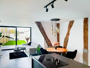 A modern living room with a sofa, coffee table, and dining table. There is a large window providing a view of the outdoors. - Charming Authentic Home With Garden (Borgloon)