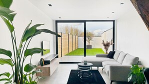 A modern living room with a grey sofa, a coffee table, and a large window overlooking a garden. - Charming Authentic Home With Garden (Borgloon)