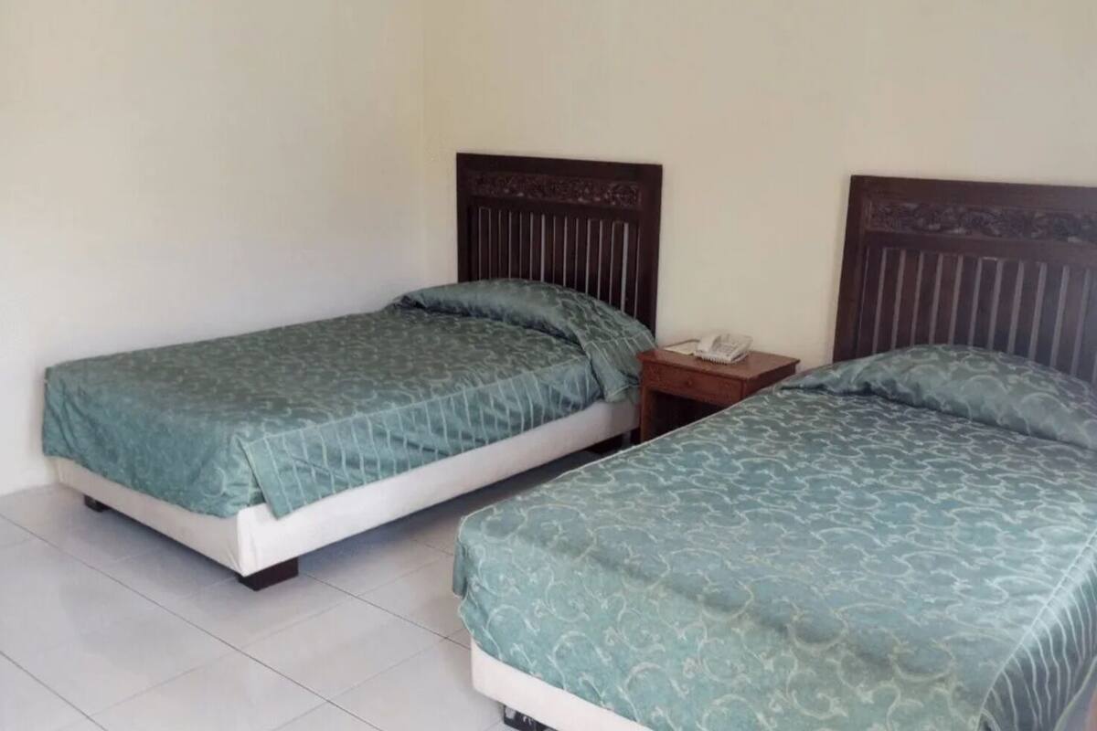 A room with two single beds, each with a wooden headboard, a small bedside table with a phone, and a light-colored tiled floor.