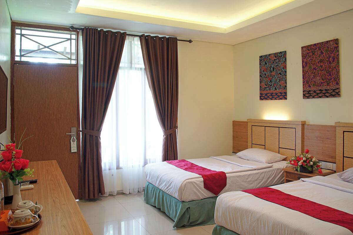 A hotel room with two beds, a wooden desk, a vase with flowers, and artwork on the wall.