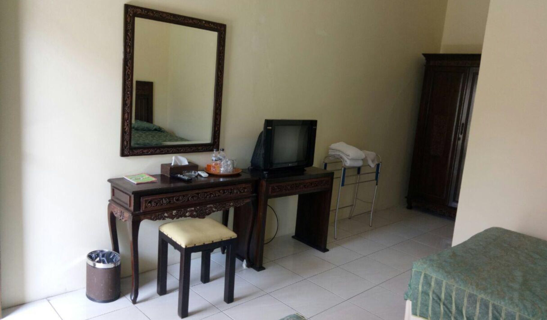 A small, simple room with a wooden desk, a television, a mirror, a small stool, a waste bin, and a bed with a green blanket.
