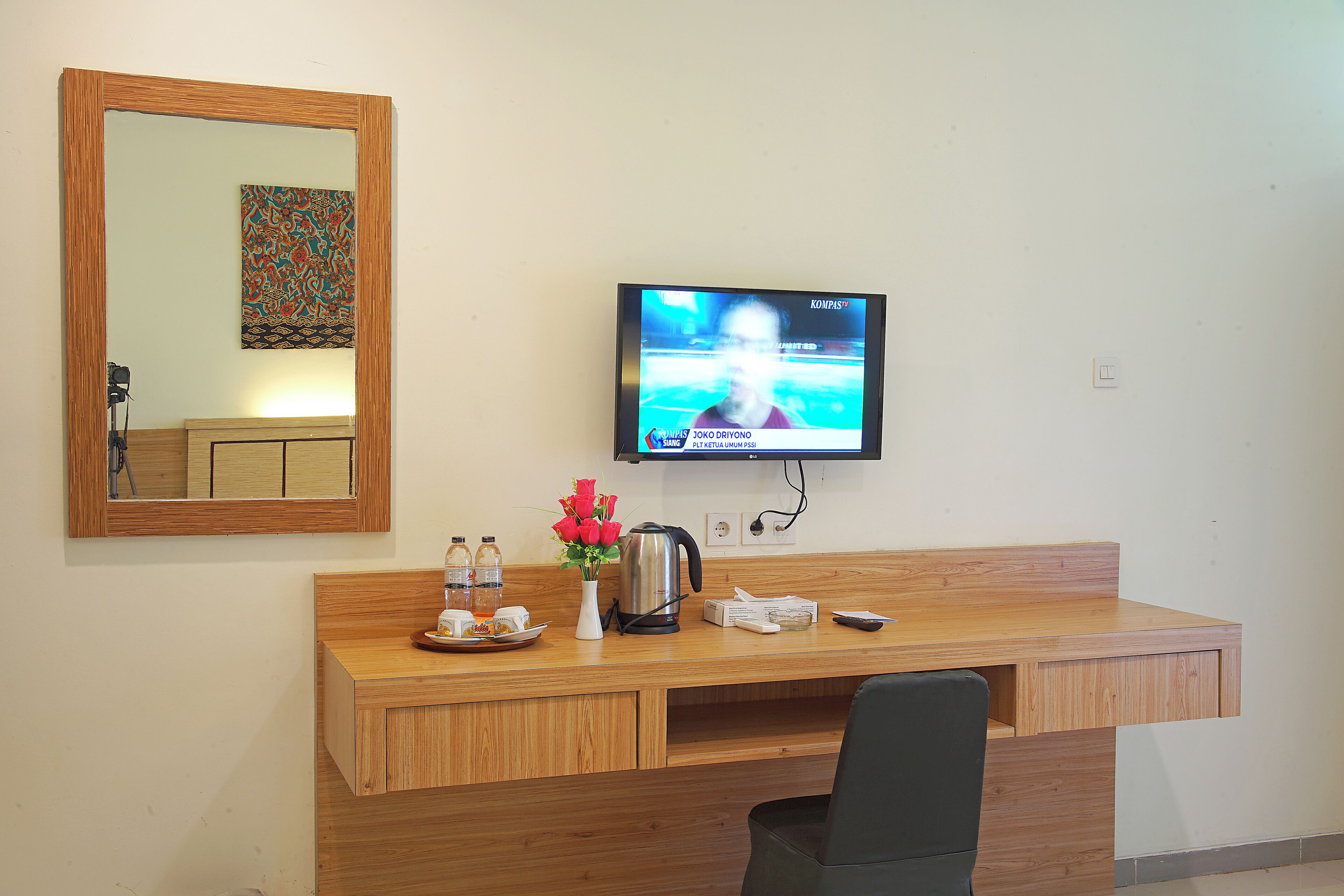 Een houten bureau met een televisie, een spiegel, een vaas met rode bloemen, een waterkoker en een afstandsbediening.