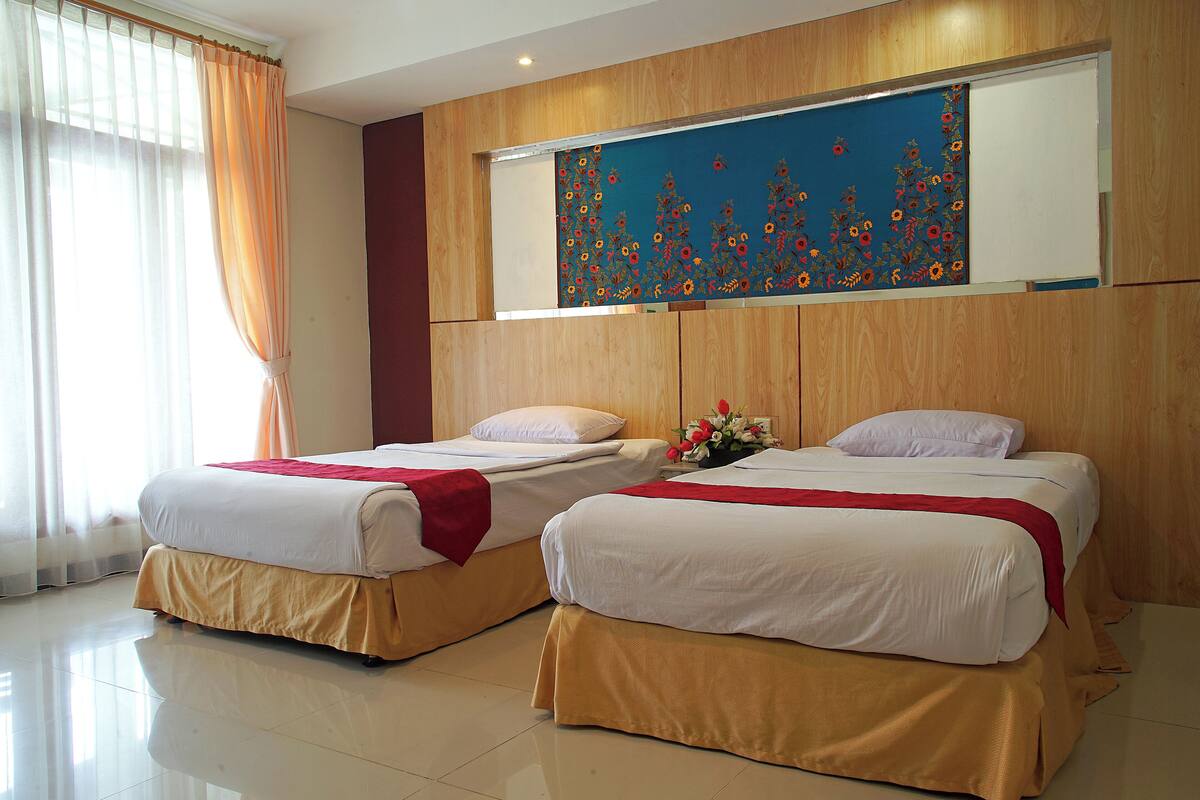 A hotel room with two beds, a wooden headboard, a floral artwork above the beds, and a window with curtains.