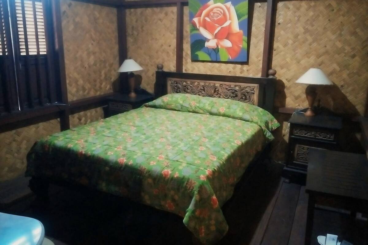 A room with a bed, two nightstands, a lamp on each, a wooden headboard, and a wall painting of a rose.