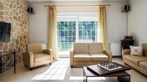 A living room with a stone wall, two beige sofas, a coffee table with a chessboard, and a large window with curtains. - Villa Nefeli with Private Pool by Konnect (Corfu)