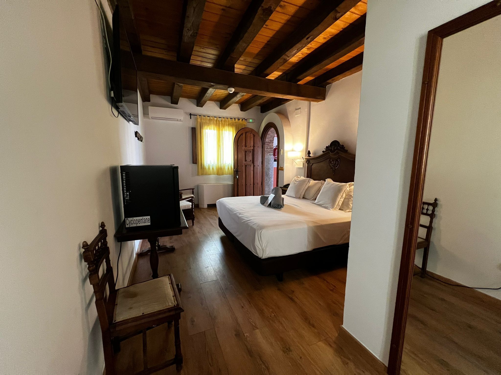 Un dormitorio con techo de madera, cama con ropa de cama blanca, televisor sobre un mueble bajo y una silla de madera.
