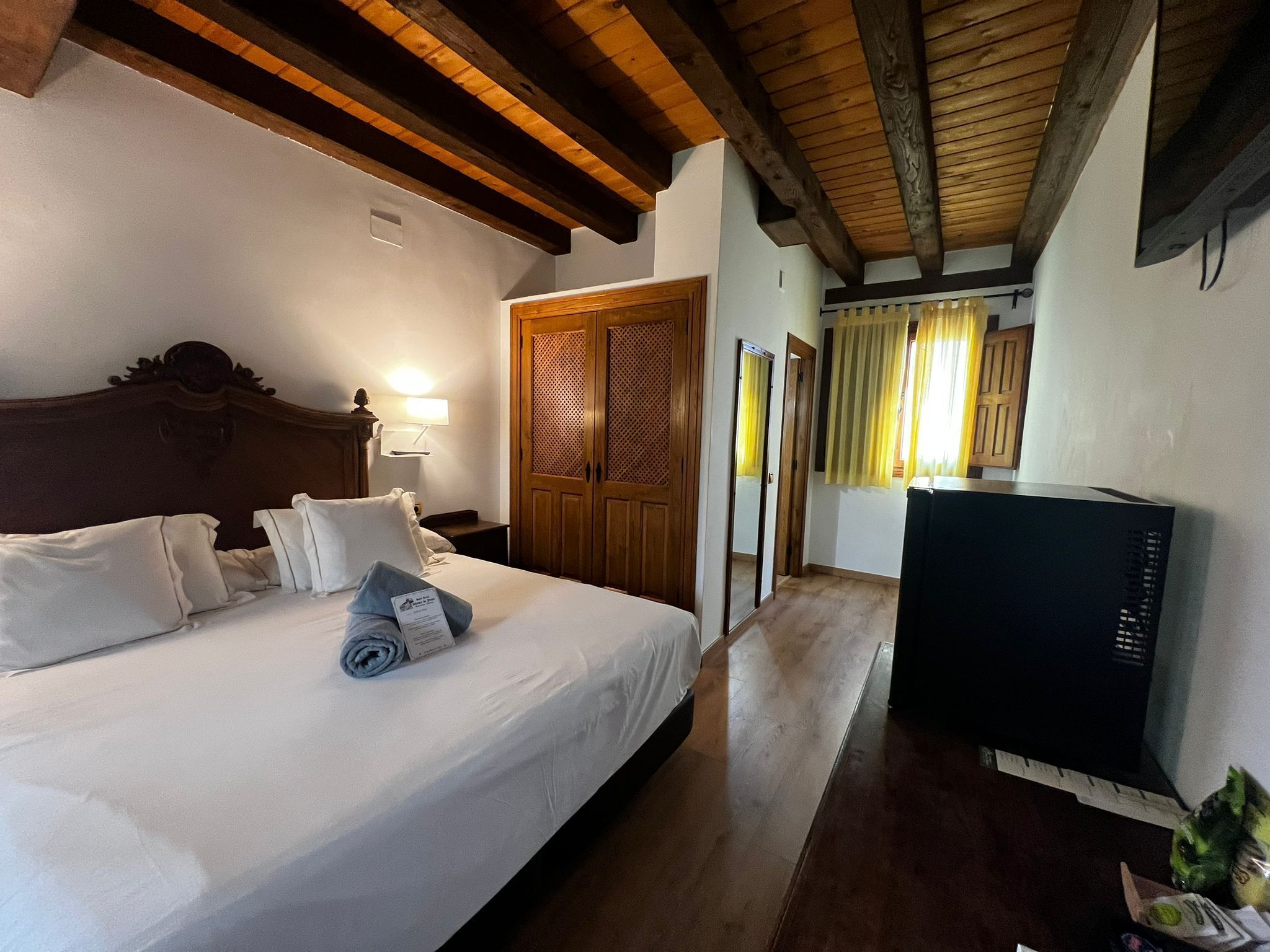 Habitación de hotel con techo de madera, cama con ropa blanca, armario de madera, televisor y una mesita con una planta.