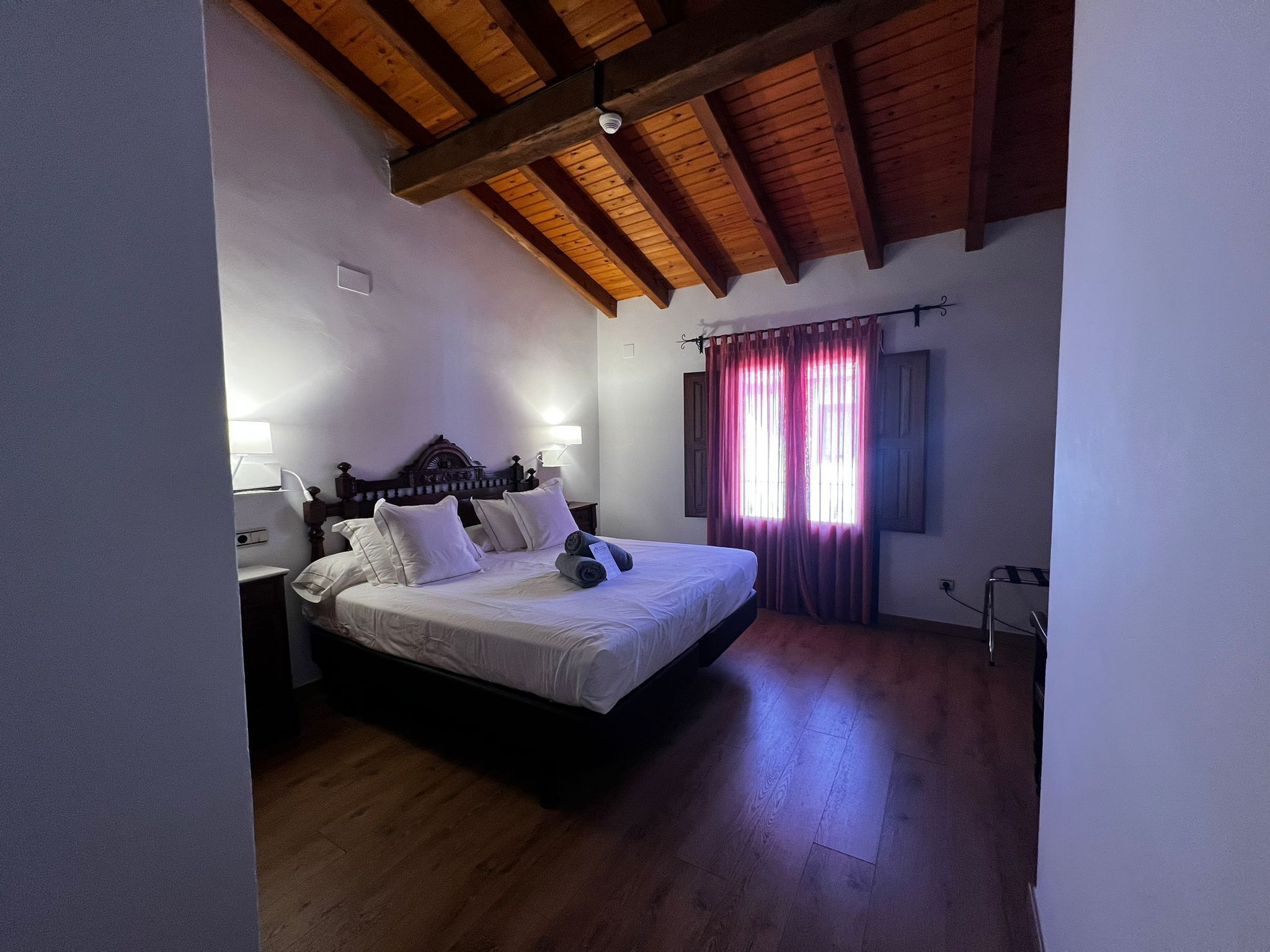 A bedroom with a wooden ceiling, a bed with white linens, a nightstand, and a window with purple curtains.