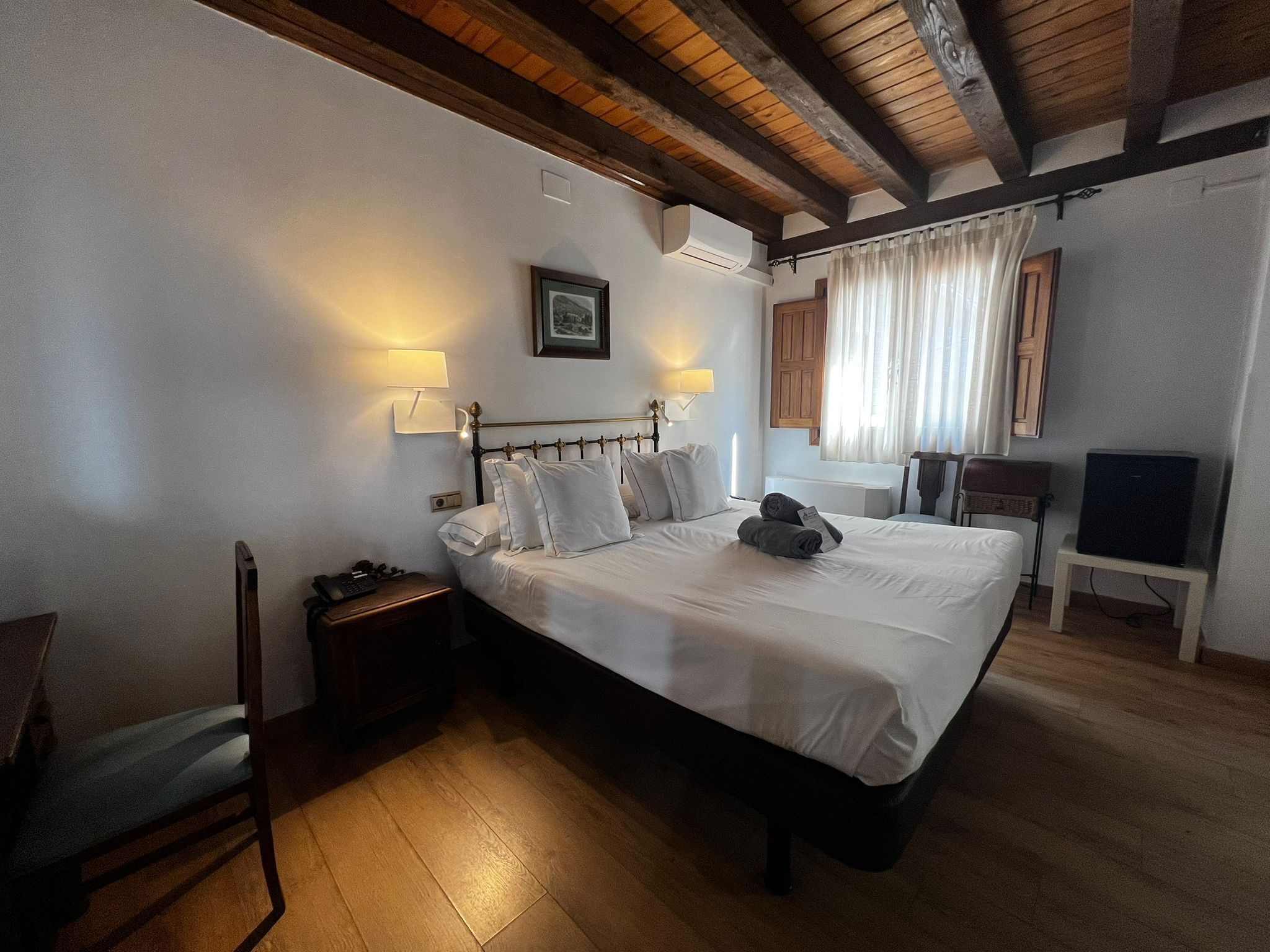 Una habitación de hotel con una cama grande, lámparas de noche, techo de madera, ventana con cortinas, un televisor y una silla.