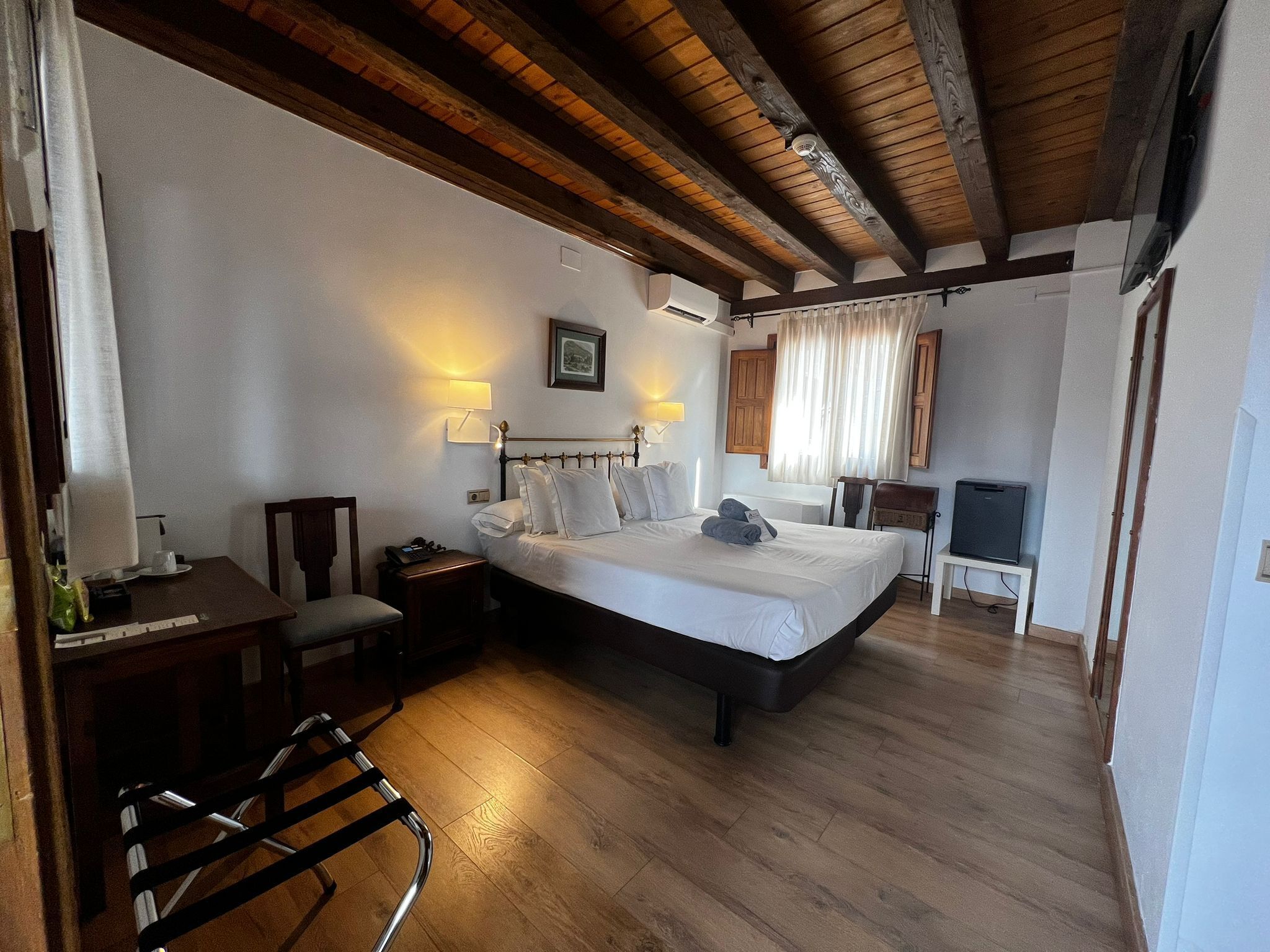 A bedroom with a wooden ceiling, a bed with white linens, a desk with a chair, and a television on a stand.