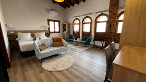 A spacious room with a bed, a sofa, a dining table, and a chair. There are arched windows and a wooden ceiling.