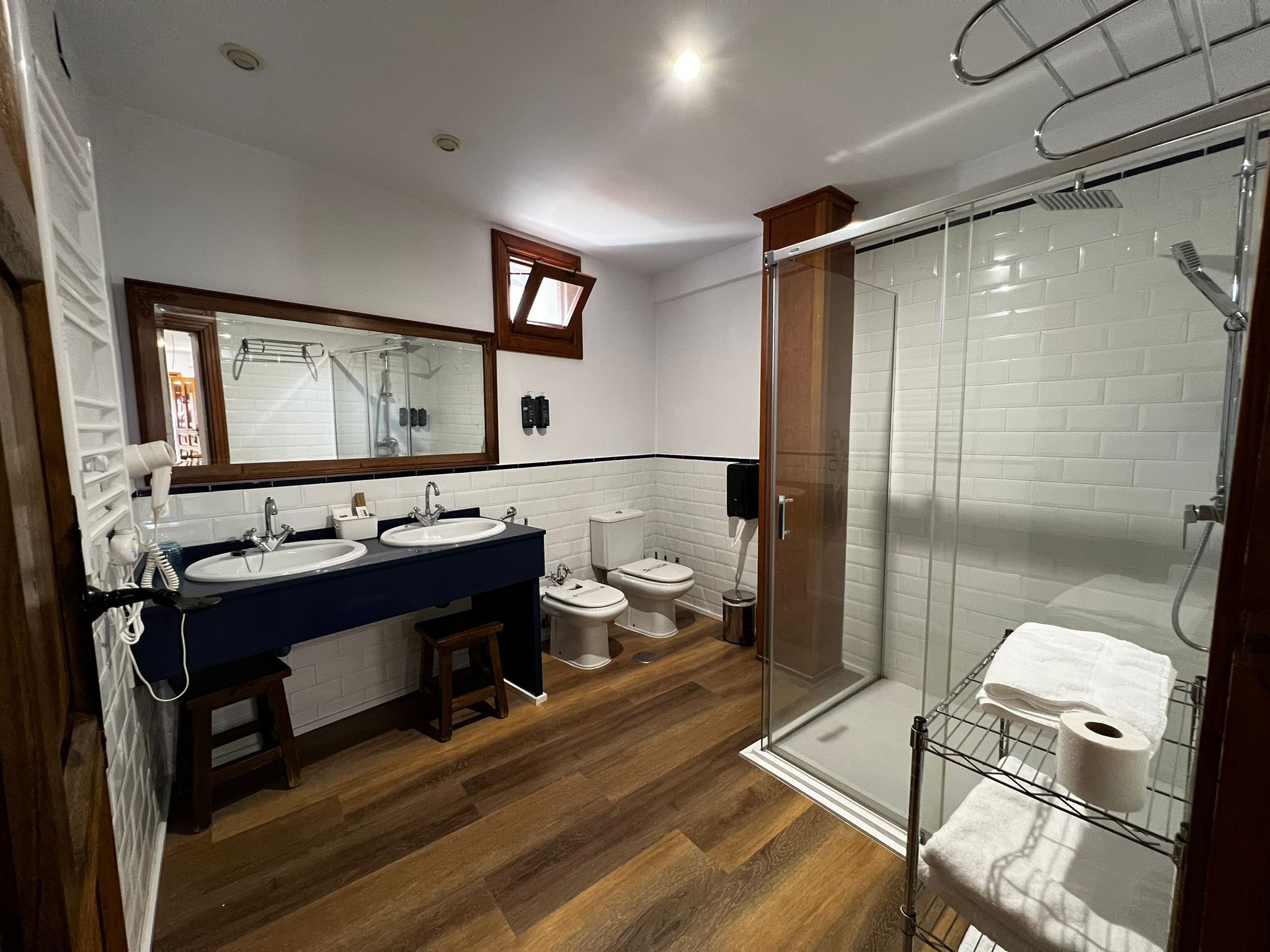 Un baño con dos lavamanos, un espejo, un inodoro y zona de ducha con mampara de vidrio.
