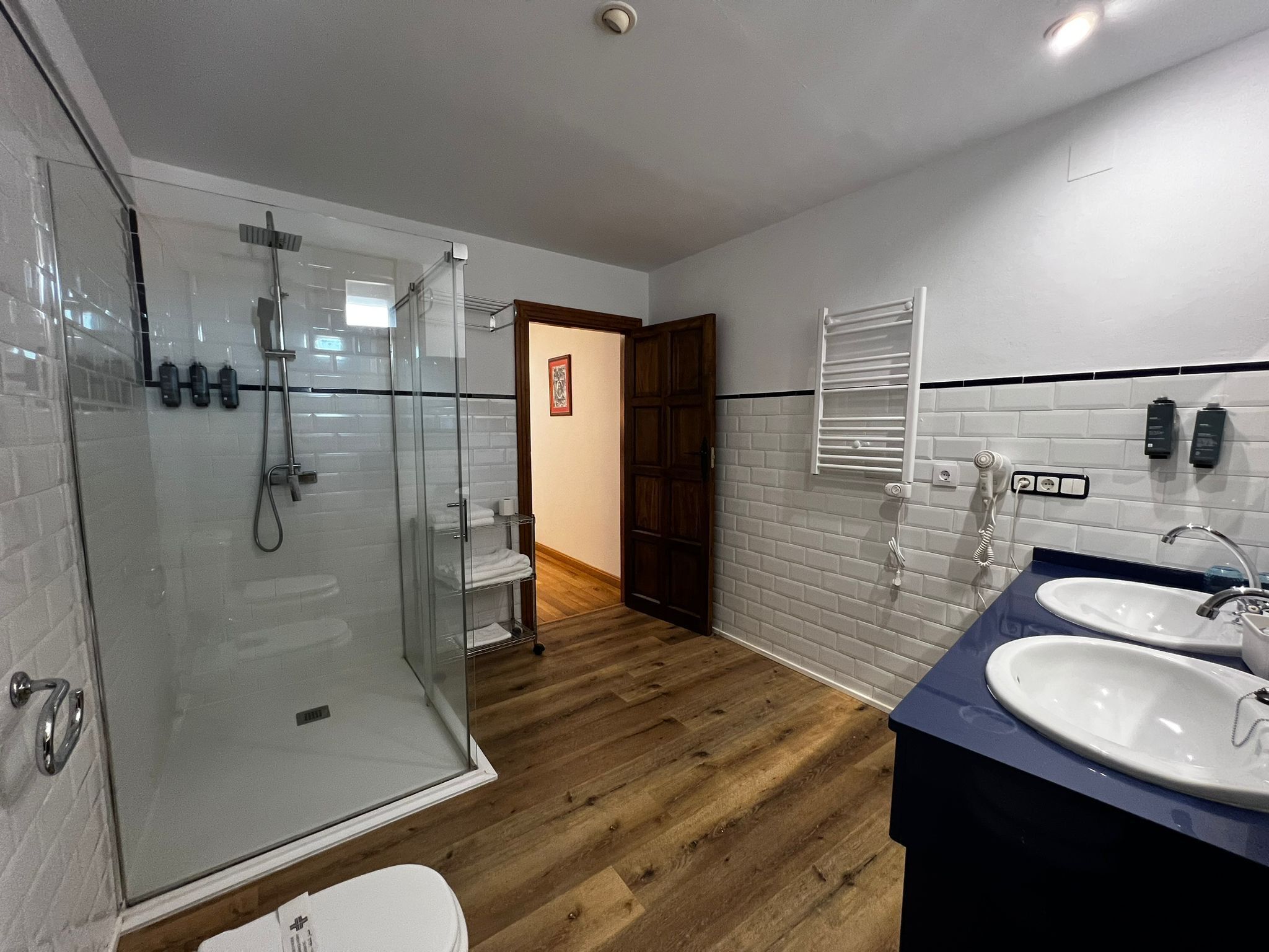 A modern bathroom with a glass shower enclosure, wooden flooring, a white toilet, and two sinks with faucets.