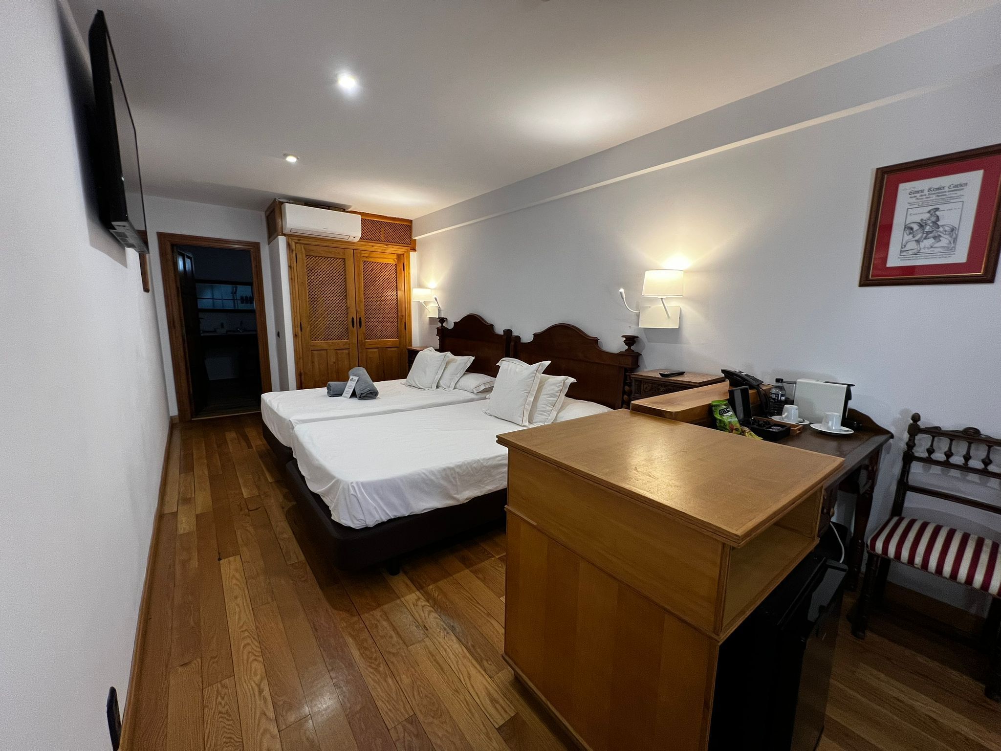 A hotel room with two beds, a wooden desk, a chair, a TV, a lamp, and a framed picture on the wall.