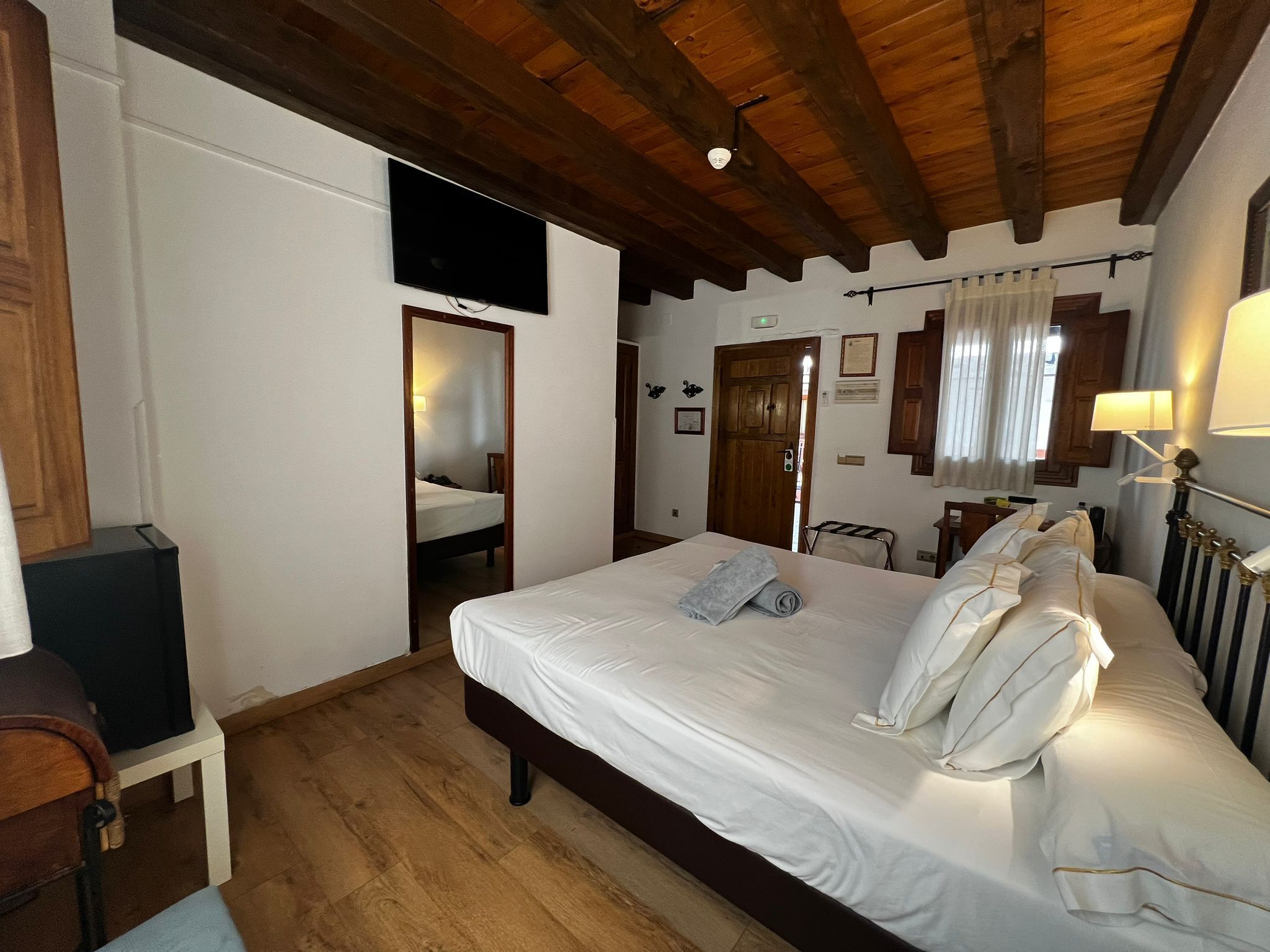 A hotel room with a wooden ceiling, a bed with white linens, a flat-screen TV mounted on the wall, and a small bedside table with a lamp.