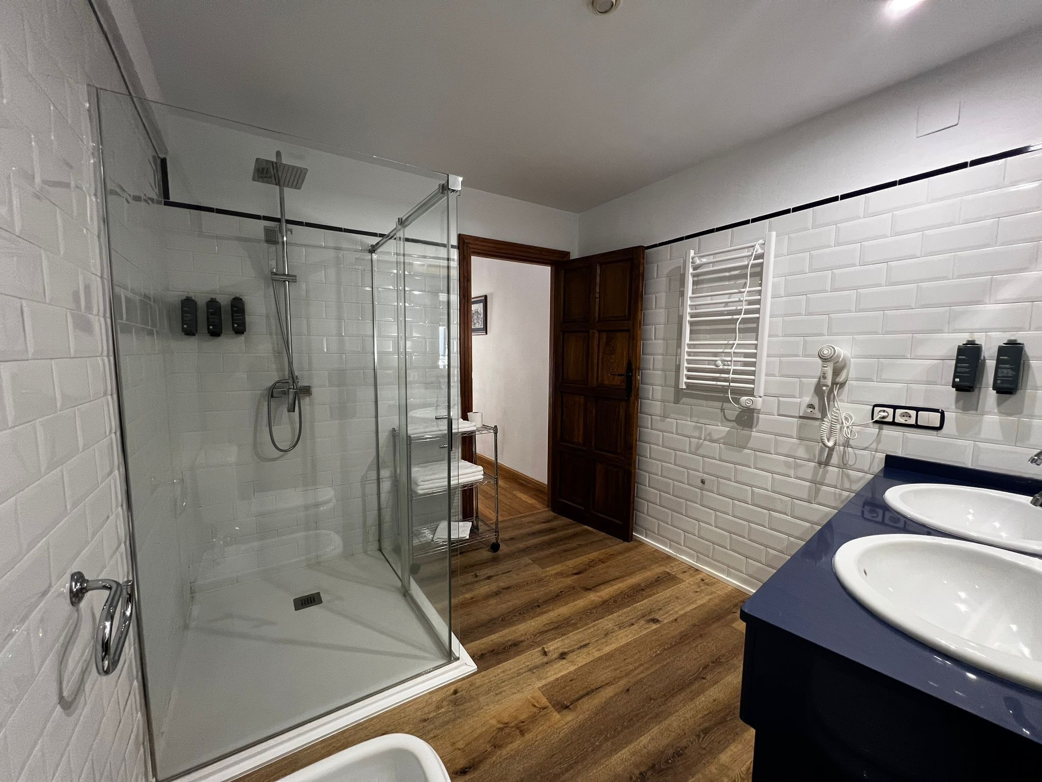 A modern bathroom with a glass-enclosed shower, a wooden floor, and a dark blue vanity with two sinks.
