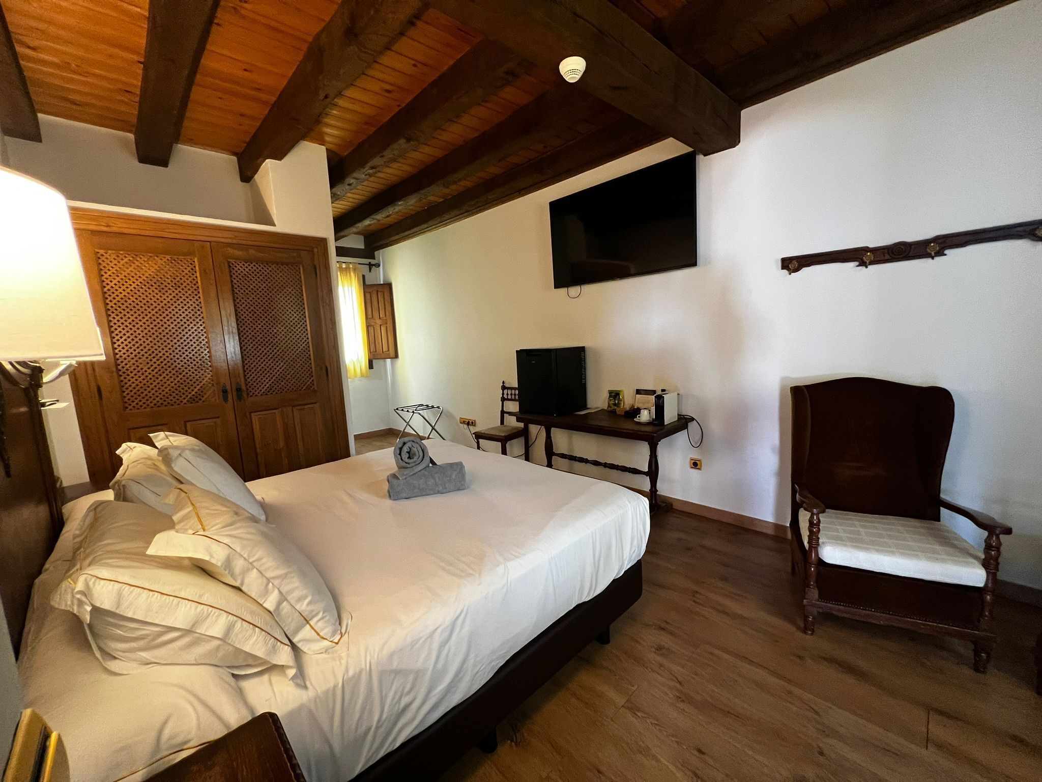 A bedroom with a wooden bed, a television mounted on the wall, a wooden chair, and a desk with a computer.