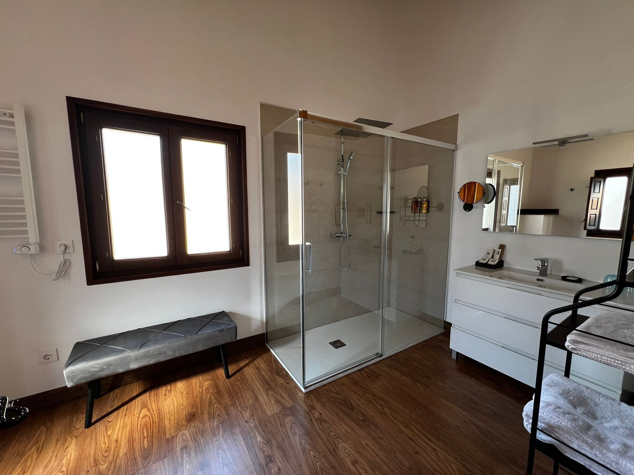 A modern bathroom with a glass shower enclosure, a bench, a vanity with a mirror, and a window providing natural light.