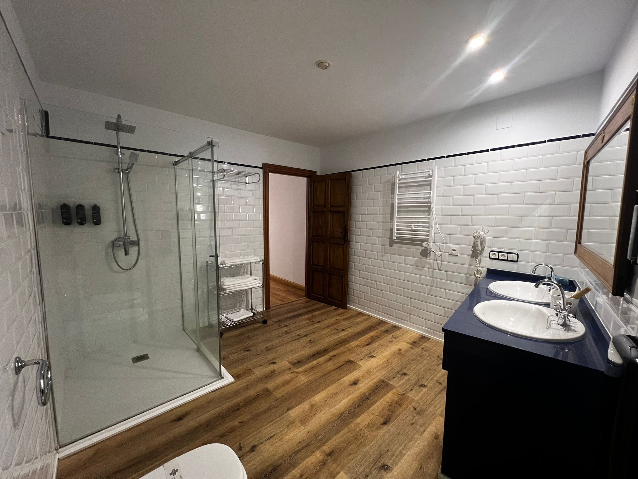 Un baño moderno con ducha envuelta en vidrio, suelo de madera, pared de azulejos blancos y un tocador oscuro con espejo.