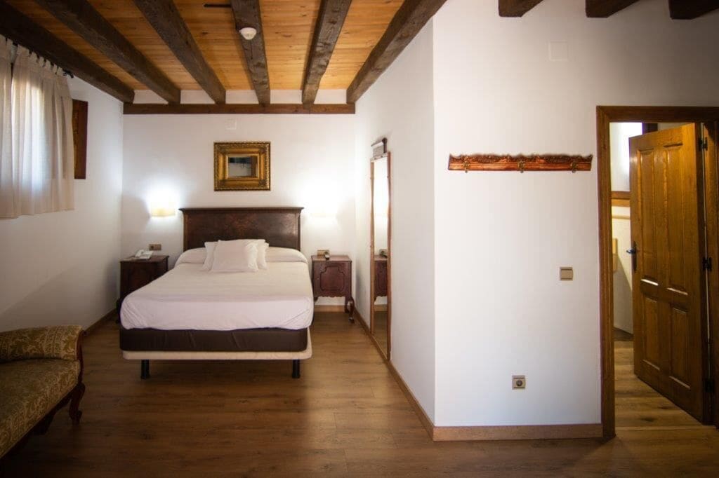 A bedroom with a wooden ceiling, a bed with white bedding, a nightstand, a bedside table, and a mirror.