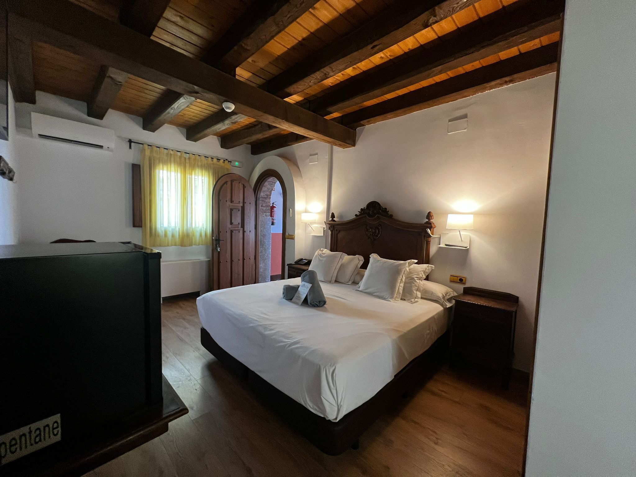 Habitación de hotel con techo de madera, cama con sábanas blancas, dos lámparas de noche y un televisor.