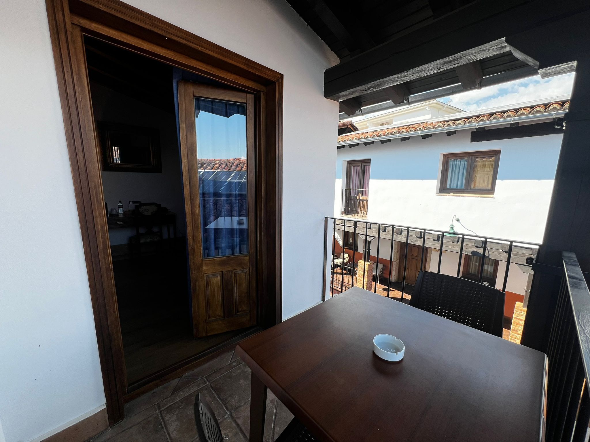 Un balcón con una mesa y silla de madera, una puerta que da a una habitación y vistas de edificios vecinos.