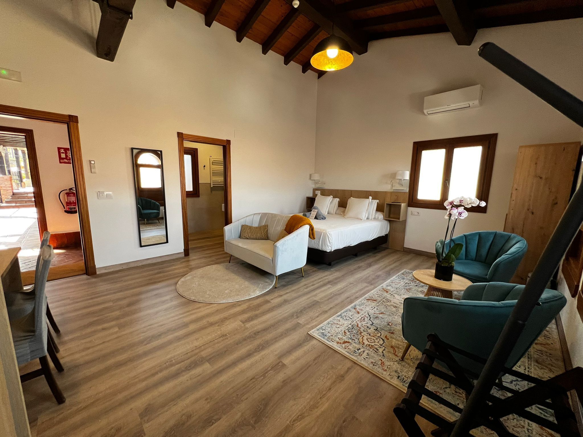 Una habitación amplia con una cama grande, una zona de estar con un sofá y sillones, una mesita con un florero y un techo de madera.